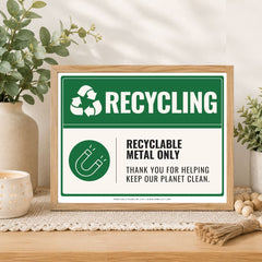 A framed recycling sign displayed on a wooden table in a styled home setting with plants, a candle, and decorative beads. The sign features a green and white design with text reading 'RECYCLING' and 'RECYCLABLE METAL ONLY'.