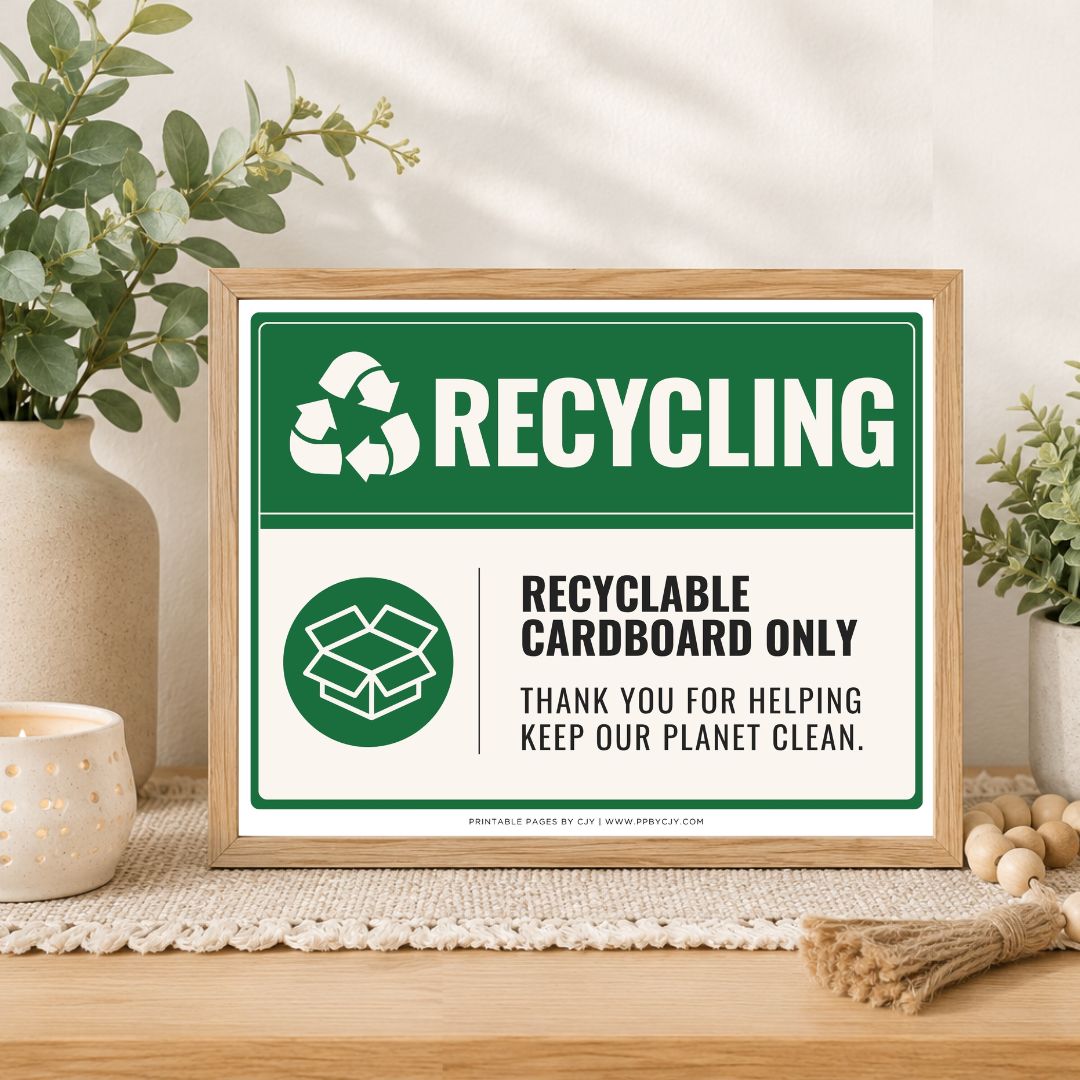 A framed recycling sign displayed on a wooden table in a styled home setting with plants and a candle. The sign features a green and white design with the text 'RECYCLING' and 'RECYCLABLE CARDBOARD ONLY'.