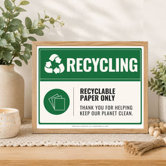 A framed recycling sign displayed on a wooden table in a styled home setting with plants, a candle, and decorative beads. The sign has a green and white design with the text 'RECYCLING' and 'RECYCLABLE PAPER ONLY'.