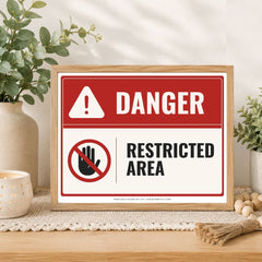 A framed warning sign with a red and white background, featuring the word 'DANGER' and a crossed-out hand symbol with the words 'RESTRICTED AREA.' The sign is displayed on a tabletop in a styled interior setting with plants, a candle, and decorative props.