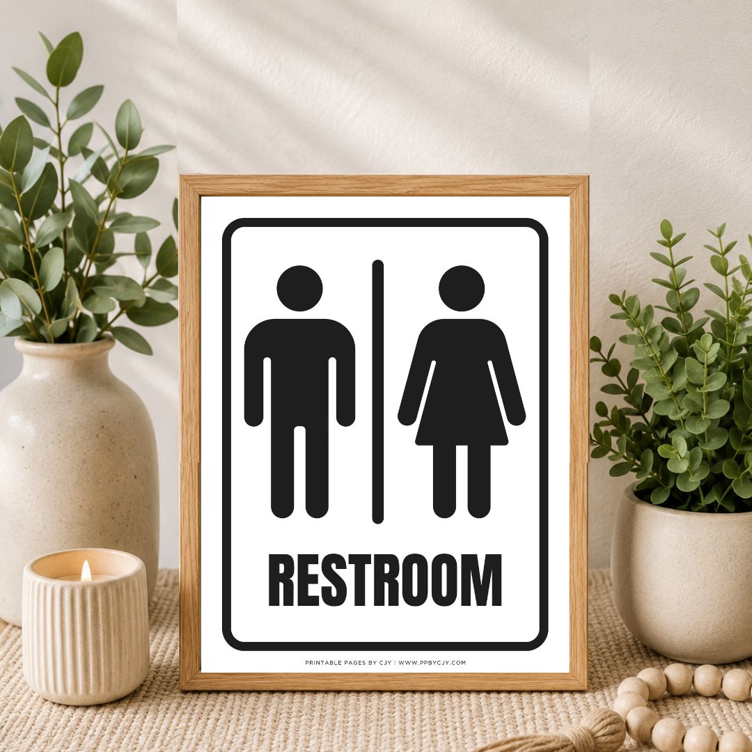 A framed restroom sign with black male and female restroom symbols and the word RESTROOM on a white background, set in a styled interior scene with plants, a candle, and a wooden frame.