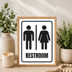 A framed restroom sign with black male and female restroom symbols and the word RESTROOM on a white background, set in a styled interior scene with plants, a candle, and a wooden frame.