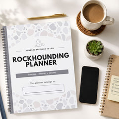 A spiral-bound 'Rockhounding Planner' notebook is shown in a styled flat-lay arrangement on a white surface, surrounded by a cup of coffee, a pen, a small succulent, a smartphone, and another notebook.