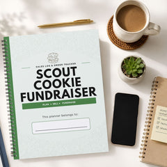 A spiral-bound planner titled 'SCOUT COOKIE FUNDRAISER' lies on a white desk, surrounded by a cup of coffee, a pen, a small succulent, a smartphone, and another notebook.