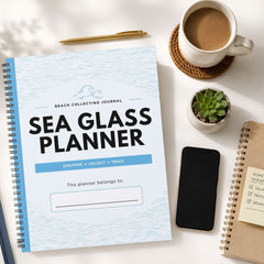 A spiral-bound Sea Glass Planner journal is shown in a styled flat-lay arrangement on a white surface, surrounded by a coffee cup, a pen, a small succulent, a smartphone, and another notebook.