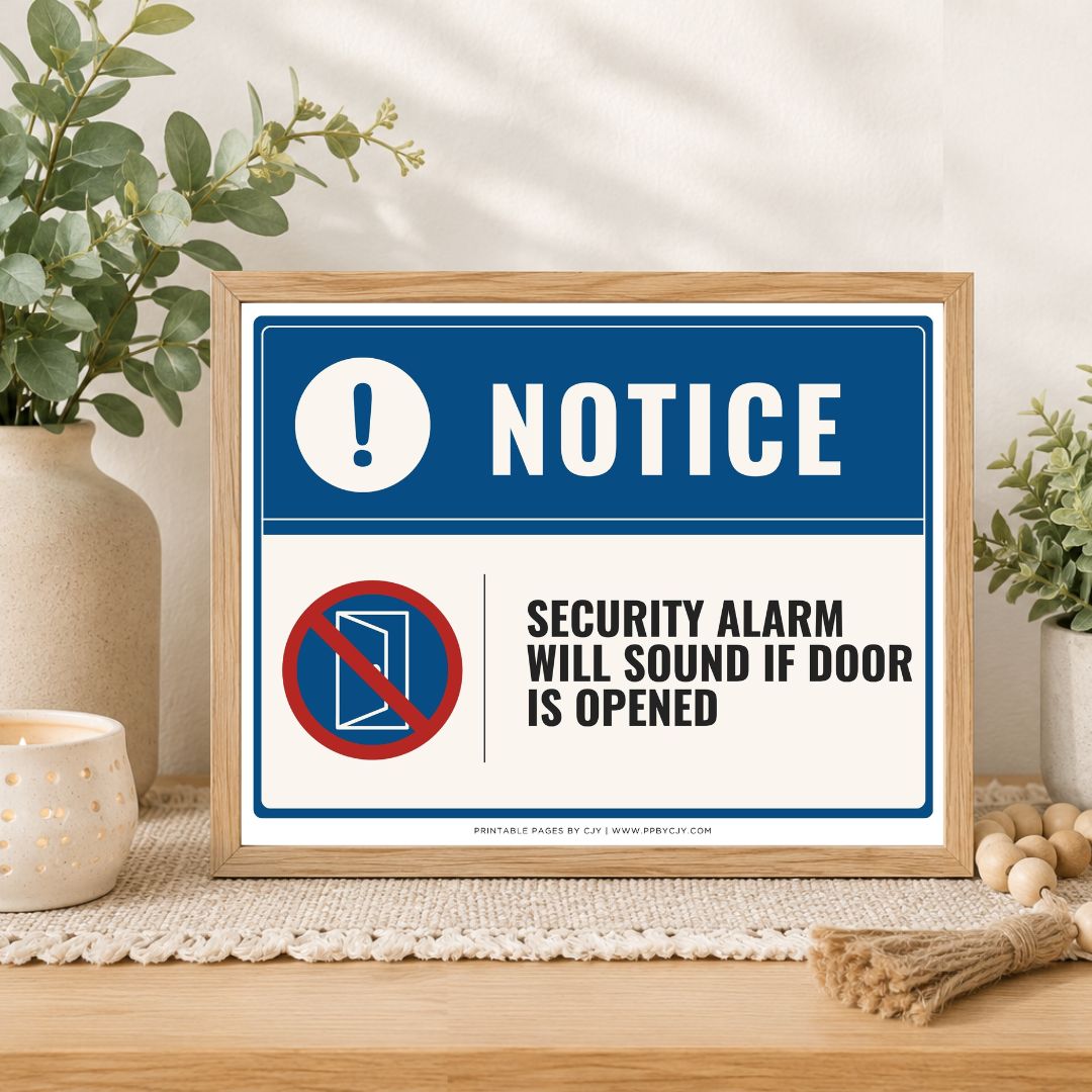 A framed printed notice sign on a tabletop in a styled interior setting. The sign has a blue header with a white exclamation mark and the word NOTICE, plus a red-and-white crossed-out door icon and the text 'SECURITY ALARM WILL SOUND IF DOOR IS OPENED.