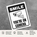 Printable Smile You're on Camera sign featuring a bold black design, security camera graphic, and surveillance notice for retail stores, offices, warehouses, parking areas, and commercial properties.