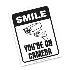 A square novelty sign with rounded corners featuring a black and white security camera graphic and the text 'SMILE YOU'RE ON CAMERA' on a white background.