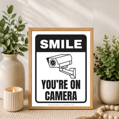 A framed novelty sign with a wood frame, featuring the text 'SMILE YOU'RE ON CAMERA' and a black-and-white security camera illustration, styled on a tabletop with plants and a candle.