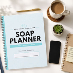 A spiral-bound 'Soap Making Planner' notebook is shown in a styled flat-lay arrangement on a white surface, surrounded by a cup of coffee, a small succulent, a smartphone, and another notebook.