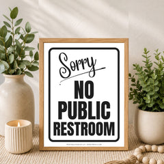 A framed novelty sign reading 'Sorry NO PUBLIC RESTROOM' in black text on a white background, styled in a home setting with potted green plants, a candle, and a wooden bead runner.
