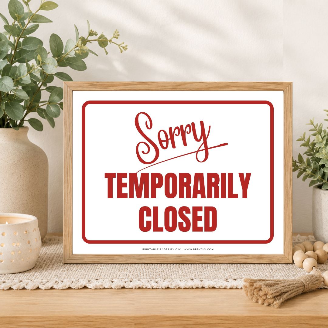 A framed sign with a light wood frame and a white background reading 'Sorry TEMPORARILY CLOSED' in red script and block lettering, styled on a tabletop with plants, a candle, and decorative beads.