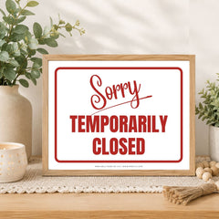 A framed sign with a light wood frame and a white background reading 'Sorry TEMPORARILY CLOSED' in red script and block lettering, styled on a tabletop with plants, a candle, and decorative beads.