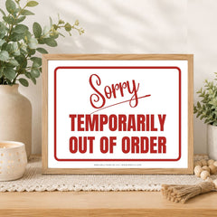 A framed typographic sign reading 'Sorry TEMPORARILY OUT OF ORDER' in red lettering on a white background, styled on a tabletop with plants, a candle, and decorative props.