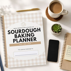 A spiral-bound Sourdough Baking Planner with a beige and white gingham cover is displayed in a styled flat-lay arrangement on a white surface, surrounded by a cup of coffee, a smartphone, a small succulent, and another notebook.