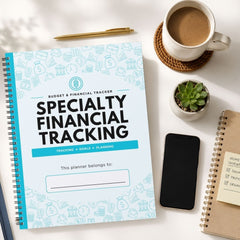 A spiral-bound planner titled 'Specialty Financial Tracking' lies on a white desk surrounded by a coffee cup, a small succulent, a smartphone, and other notebooks.