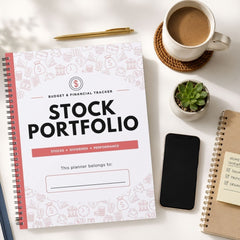 A spiral-bound stock portfolio planner notebook with a white cover and pink financial icons, styled in a flat-lay arrangement on a white desk alongside a cup of coffee, a pen, a small succulent, a smartphone, and another notebook.