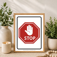 A framed art print featuring a red octagonal stop sign with a white hand symbol and the word 'STOP' on a white background, styled in a home decor setting with potted plants and a candle.