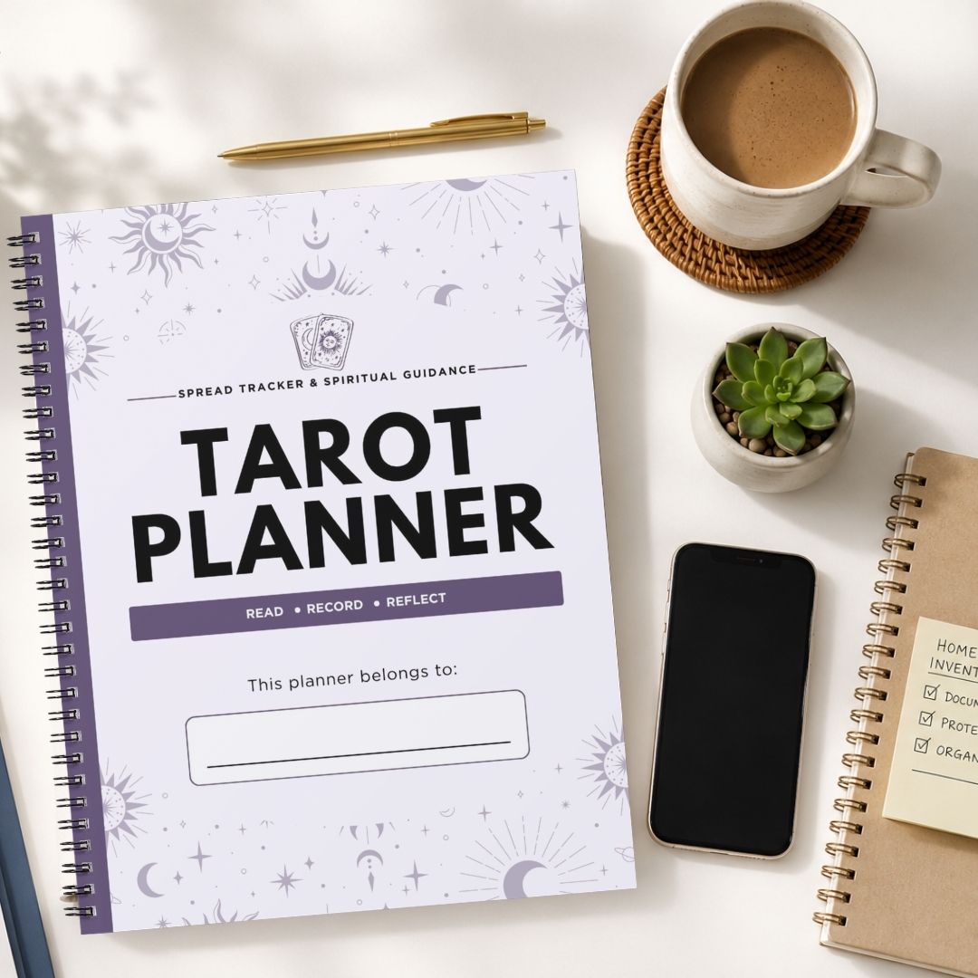 A spiral-bound tarot planner lies on a light tabletop, surrounded by a coffee cup on a coaster, a small potted succulent, a smartphone, and another notebook. The planner cover is white with black and purple text and subtle celestial line illustrations.