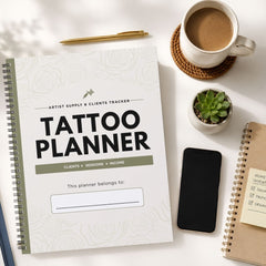 A spiral-bound 'Tattoo Planner' notebook is shown in a styled flat-lay arrangement on a white surface, alongside a cup of coffee, a pen, a small succulent, a smartphone, and another notebook.