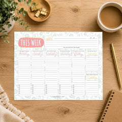 A weekly planner notepad sheet laid flat on a wooden desk, surrounded by a coffee cup, plant, binder clips, pen, and notebook. The planner has a pale floral border and sections labeled Monday through Sunday, with a pink title bubble reading THIS WEEK.