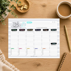 A weekly planner notepad sheet laid flat on a wooden desk, surrounded by a coffee cup, notebook, pen, and small plant. The notepad has sections for notes, monthly overview, and daily planning from Monday to Sunday.
