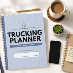 A spiral-bound trucking planner with a light blue cover is displayed in a styled flat-lay arrangement on a white desk, surrounded by a cup of coffee, a smartphone, a small succulent, and other notebooks.