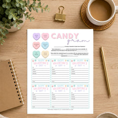 A Valentine's Day printable card set titled 'Candy from' laid flat on a wooden desk, surrounded by a coffee cup, binder clip, plant, notebook, and pen.