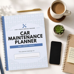 A spiral-bound Car Maintenance Planner with a white cover and blue accents is shown in a styled flat-lay arrangement on a light surface, surrounded by a coffee cup, a pen, a small succulent, a smartphone, and another notebook.