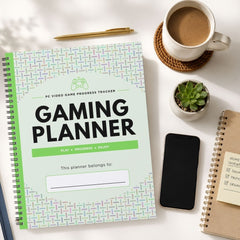 A spiral-bound gaming planner titled 'GAMING PLANNER' lies on a white tabletop, surrounded by a coffee cup in a mug, a small succulent, a smartphone, and another notebook. The planner cover has a pale mint background with a subtle geometric pattern and bold black text.