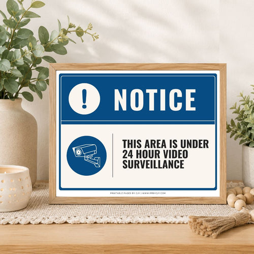 A framed notice sign on a tabletop in a styled interior setting. The sign reads 'NOTICE' with a blue warning icon and the text 'THIS AREA IS UNDER 24 HOUR VIDEO SURVEILLANCE.
