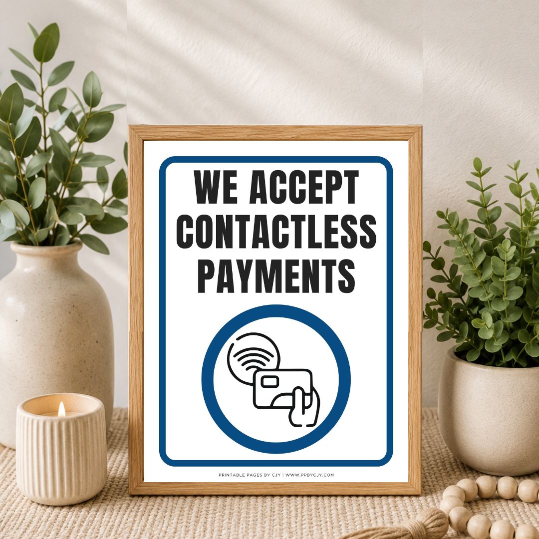 A framed sign reading 'WE ACCEPT CONTACTLESS PAYMENTS' with a blue-and-white payment icon, styled on a tabletop between two potted plants and a candle.